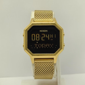 May include: Gold-toned Nixon digital watch with a mesh band. The watch face displays the time as 08:24 AM and the word "TODAY". The watch has a geometric octagonal shape and gold-colored buttons on the sides.