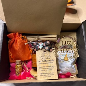 May include: A gift box filled with various items, including a small orange drawstring bag, a jar with a decorative ribbon, and a pouch with the text "LUZ ALMA". The box contains pink and red decorative elements.