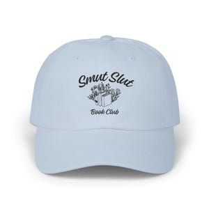 May include: Light blue baseball cap with the text "Smut Slut Book Club" in black script. The design includes two books and floral accents. The cap has a curved brim and a rounded crown.