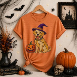 Puede incluir: Camiseta naranja con un perro de dibujos animados que lleva un sombrero de bruja morado y un collar colorido, sentado junto a una calabaza de Halloween. La camiseta forma parte de un tema de Halloween, con calabazas y una calavera en el fondo.