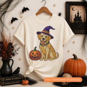 Puede incluir: Camiseta color crema con un golden retriever con un sombrero de bruja morado y un collar decorativo, con una calabaza de Halloween. El diseño es perfecto para Halloween. La camiseta está en una percha de madera.
