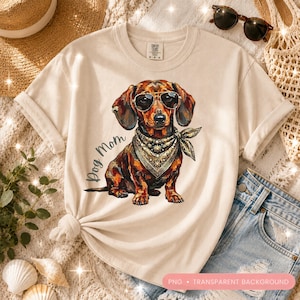 Op de afbeelding: Crèmekleurig T-shirt met een teckel met een zonnebril en een bandana. De afbeelding bevat de tekst "Dog Mom". De hond is bruin en zwart met gouden accenten. Het shirt heeft opgerolde mouwen en een geknoopte zoom.