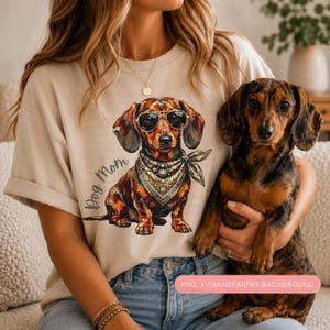 Op de afbeelding: Beige T-shirt met een afbeelding van een teckel met een zonnebril en een bandana. De tekst "Dog Mom" staat op het shirt. Een echte teckel wordt in de armen van de persoon gehouden.
