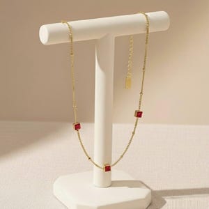 Peut inclure: Un collier doré avec quatre pierres rouges carrées. Le collier est présenté sur un présentoir à bijoux blanc sur fond neutre. La chaîne est délicate avec de petites perles rondes.
