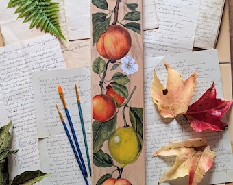 May include: Wooden chopping board with a painted fruit design. The design features peaches, apples, and a lemon on a branch with green leaves and white flowers. Two paintbrushes are visible. The board has a hole at the top.