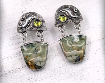 May include: A pair of handcrafted silver earrings. Each earring features a textured, half-circle top with a small, round, yellow-green stone and a curved silver accent. A larger, shield-shaped, green and brown stone hangs below, connected by a silver chain.