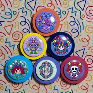 May include: Seven colourful, round pinback buttons with distinct cartoon designs. Each button showcases a different illustration, such as a purple monster, a smiling bunny, and a sad jester. The buttons are displayed on a patterned fabric background.