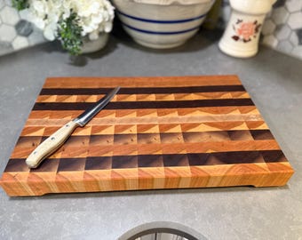 May include: A rectangular wooden cutting board with a checkerboard pattern of light and dark wood. A stainless steel knife with a light-colored handle rests on the board. The board is approximately 16 inches long and 12 inches wide.