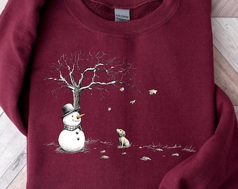 Puede incluir: Sudadera burdeos con un estampado de escena invernal. El diseño presenta un muñeco de nieve con sombrero de copa y bufanda, un árbol y un perro pequeño. El perro mira hacia el árbol. La sudadera es de color burdeos oscuro.