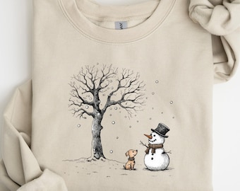 Puede incluir: Sudadera beige con una ilustración de escena invernal. El diseño muestra un árbol desnudo, un muñeco de nieve con sombrero de copa y bufanda, y un perro pequeño. Los copos de nieve caen a su alrededor. La sudadera tiene mangas largas y cuello redondo.