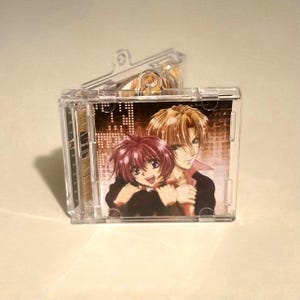 Może przedstawiać: Przezroczyste plastikowe etui na CD z grafiką anime przedstawiającą dwie postacie obejmujące się. Grafika ma tło w odcieniach sepii z krajobrazem miejskim. Postacie mają rude i blond włosy. Etui jest przezroczyste.