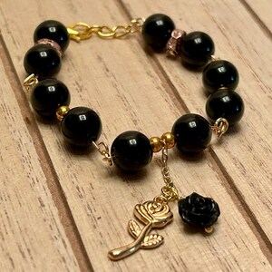 May include: A black beaded bracelet with gold-coloured accents and a rose charm. The bracelet features large, round black beads connected by gold-coloured chain links. A gold-coloured rose charm and a black rose charm dangle from the bracelet.