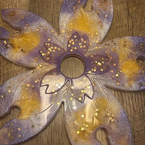 May include: A flower-shaped resin coaster with a central hole and cutouts for glasses. The coaster has a mix of purple, gold, and white colours, with gold star-shaped glitter accents. Designed to hold multiple glasses, it's a decorative item.