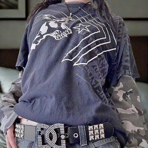 Puede incluir: Una camiseta gráfica gris oscuro con un diseño de calavera y estrella, superpuesta a una camiseta de manga larga de camuflaje. El atuendo incluye un cinturón negro con tachuelas y una hebilla grande adornada. El estilo general es atrevido e informal.