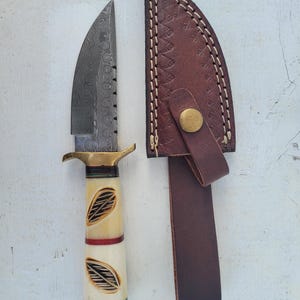 May include: A Damascus steel knife with a cream-colored handle featuring leaf designs and a brown leather sheath. The knife has a brass bolster and a curved blade. The sheath has a snap closure and a belt loop.