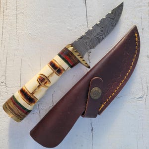 May include: A Damascus steel knife with a bone handle and a brown leather sheath. The handle features a decorative design with red, green, and brown accents. The sheath has a snap closure with a decorative emblem.