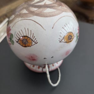 May include: A spherical white ceramic ornament featuring a hand-painted face with large orange eyes, long eyelashes, and pink cheeks. The top has brown swirling hair patterns, and the base is decorated with pink and white stripes and a hanging string.