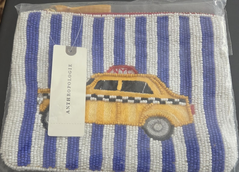 May include: Beaded pouch with a yellow taxi cab design on a blue and white striped background. The pouch has a zipper closure and a tag that says Anthropologie.