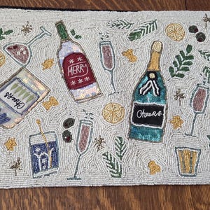 May include: A rectangular table runner featuring intricate beadwork on a white background. The design displays various cocktail bottles, champagne flutes, and glasses with text including "Fizz the Season," "Merry," and "Cheers" in colorful beads.