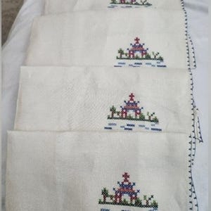 May include: A set of four off-white rectangular fabric napkins, each featuring an identical cross-stitch embroidery of a small pagoda structure in red, blue, and green thread, with a dark blue decorative stitched border along the side edge.