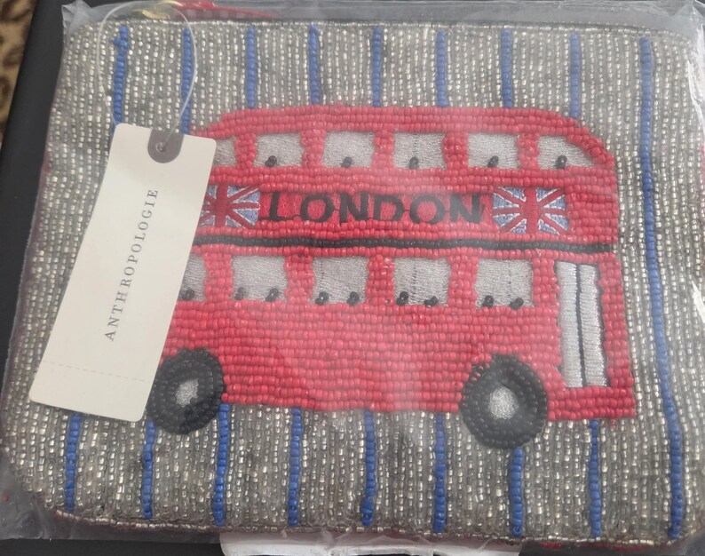 May include: A beaded clutch featuring a red double-decker bus design with the word "LONDON" and Union Jack flags. The bag has a silver and blue striped background and a tag that reads "ANTHROPOLOGIE".