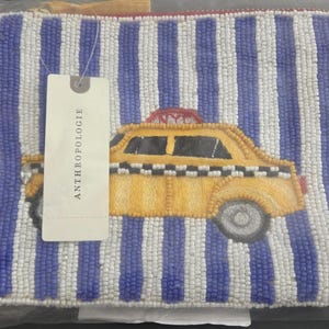 May include: Beaded clutch with a vertical striped pattern in white and blue. A yellow taxi cab design is embroidered on the front. The tag reads ANTHROPOLOGIE.