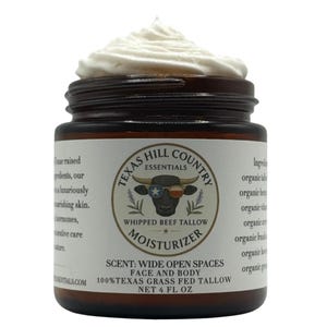 Puede incluir: Un tarro de crema hidratante de sebo de res batida de Texas Hill Country Essentials. El tarro es marrón oscuro con una etiqueta blanca con un toro con gafas de sol. El tarro contiene una crema hidratante blanca batida. La etiqueta dice "Moisturizer" y "118 ml netos".