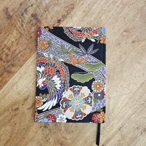 Puede incluir: Un cuaderno rectangular con una cubierta negra con un dise&ntilde;o floral y de p&aacute;jaros colorido. El dise&ntilde;o incluye flores naranjas, rojas, azules y blancas, junto con un p&aacute;jaro y hojas verdes. Se adjunta un marcador de cinta negra.