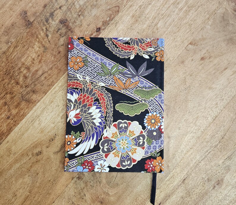 Puede incluir: Un cuaderno rectangular con una cubierta negra con un dise&ntilde;o floral y de p&aacute;jaros colorido. El dise&ntilde;o incluye flores naranjas, rojas, azules y blancas, junto con un p&aacute;jaro y hojas verdes. Se adjunta un marcador de cinta negra.