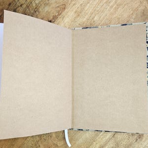 K&ouml;nnte beinhalten: Ein offenes Notizbuch mit leeren, beigefarbenen Seiten und einem wei&szlig;en Band-Lesezeichen. Der Einband hat ein Muster in Schwarz-, Grau- und Beiget&ouml;nen. Das Notizbuch liegt auf einer Holzoberfl&auml;che.