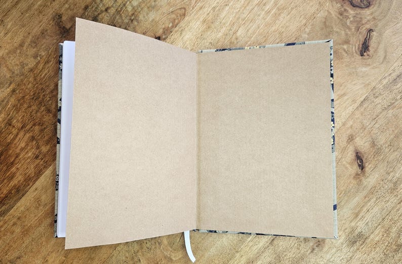 K&ouml;nnte beinhalten: Ein offenes Notizbuch mit leeren, beigefarbenen Seiten und einem wei&szlig;en Band-Lesezeichen. Der Einband hat ein Muster in Schwarz-, Grau- und Beiget&ouml;nen. Das Notizbuch liegt auf einer Holzoberfl&auml;che.