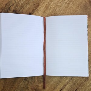 Puede incluir: Cuaderno abierto con p&aacute;ginas rayadas y un marcap&aacute;ginas de cinta marr&oacute;n. El cuaderno est&aacute; sobre una superficie de madera. Las p&aacute;ginas son blancas con l&iacute;neas horizontales. El marcap&aacute;ginas es una cinta fina de color marr&oacute;n oscuro.