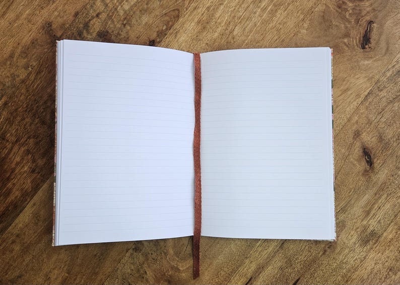 Puede incluir: Cuaderno abierto con p&aacute;ginas rayadas y un marcap&aacute;ginas de cinta marr&oacute;n. El cuaderno est&aacute; sobre una superficie de madera. Las p&aacute;ginas son blancas con l&iacute;neas horizontales. El marcap&aacute;ginas es una cinta fina de color marr&oacute;n oscuro.