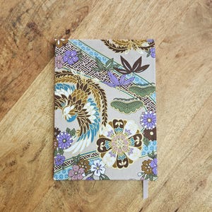 May include: A rectangular notebook with a beige cover featuring a colorful floral and bird design. The design includes shades of brown, blue, purple, and white. A light pink ribbon bookmark is visible.