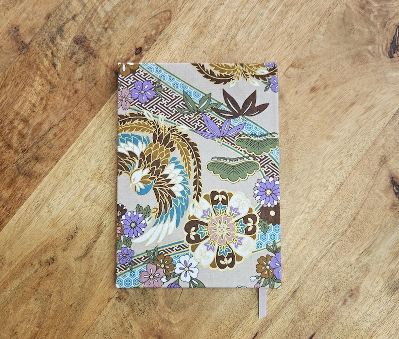 May include: A rectangular notebook with a beige cover featuring a colorful floral and bird design. The design includes shades of brown, blue, purple, and white. A light pink ribbon bookmark is visible.