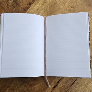 May include: An open notebook with lined pages and a light brown ribbon bookmark. The notebook's cover has a patterned design. The notebook is on a wooden surface.