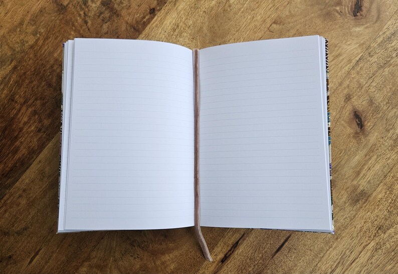 May include: An open notebook with lined pages and a light brown ribbon bookmark. The notebook's cover has a patterned design. The notebook is on a wooden surface.