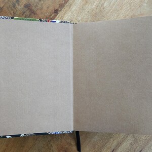Puede incluir: Un cuaderno abierto con p&aacute;ginas en blanco de color marr&oacute;n claro. La cubierta y los bordes presentan un colorido estampado floral sobre un fondo negro. Se ve una cinta negra como marcap&aacute;ginas. El cuaderno est&aacute; sobre una superficie de madera.
