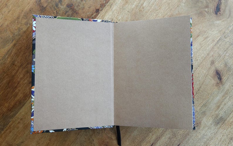 Puede incluir: Un cuaderno abierto con p&aacute;ginas en blanco de color marr&oacute;n claro. La cubierta y los bordes presentan un colorido estampado floral sobre un fondo negro. Se ve una cinta negra como marcap&aacute;ginas. El cuaderno est&aacute; sobre una superficie de madera.