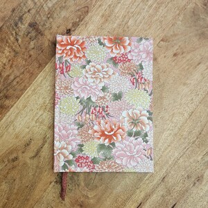 Puede incluir: Un cuaderno con un estampado floral en tonos rosa, naranja y amarillo sobre un fondo rosa. Cuenta con una cinta marr&oacute;n como marcap&aacute;ginas. El cuaderno est&aacute; sobre una superficie de madera.