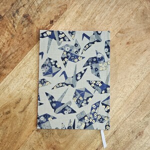 K&ouml;nnte beinhalten: Ein rechteckiges Notizbuch mit einem gemusterten Einband mit Origami-Kranichen in Blau- und Wei&szlig;t&ouml;nen auf hellgrauem Hintergrund. Ein wei&szlig;es Lesezeichenband ist sichtbar. Das Notizbuch liegt auf einer Holzoberfl&auml;che.