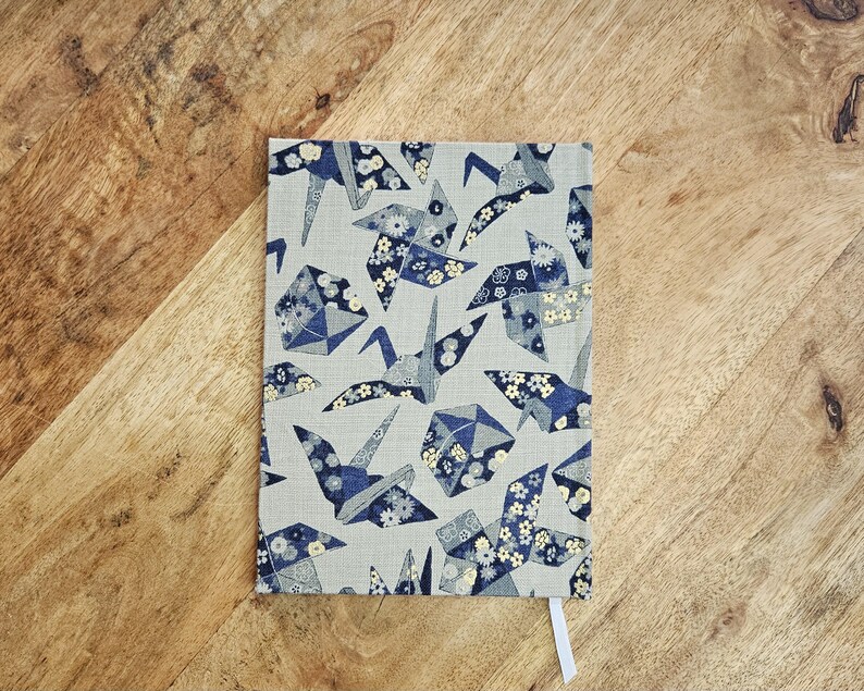K&ouml;nnte beinhalten: Ein rechteckiges Notizbuch mit einem gemusterten Einband mit Origami-Kranichen in Blau- und Wei&szlig;t&ouml;nen auf hellgrauem Hintergrund. Ein wei&szlig;es Lesezeichenband ist sichtbar. Das Notizbuch liegt auf einer Holzoberfl&auml;che.