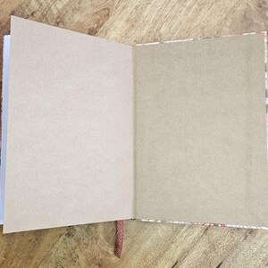 Puede incluir: Un diario abierto con p&aacute;ginas en blanco de color marr&oacute;n claro. El diario tiene un borde decorativo estampado y un marcador de cinta roja. El diario est&aacute; sobre una superficie de madera.