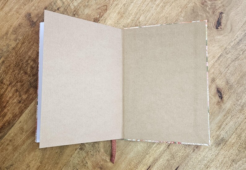 Puede incluir: Un diario abierto con p&aacute;ginas en blanco de color marr&oacute;n claro. El diario tiene un borde decorativo estampado y un marcador de cinta roja. El diario est&aacute; sobre una superficie de madera.