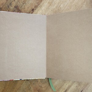 May include: An open notebook with blank, tan-colored pages. The notebook's binding has a colorful floral pattern. A green ribbon bookmark is visible. The notebook is on a wooden surface.