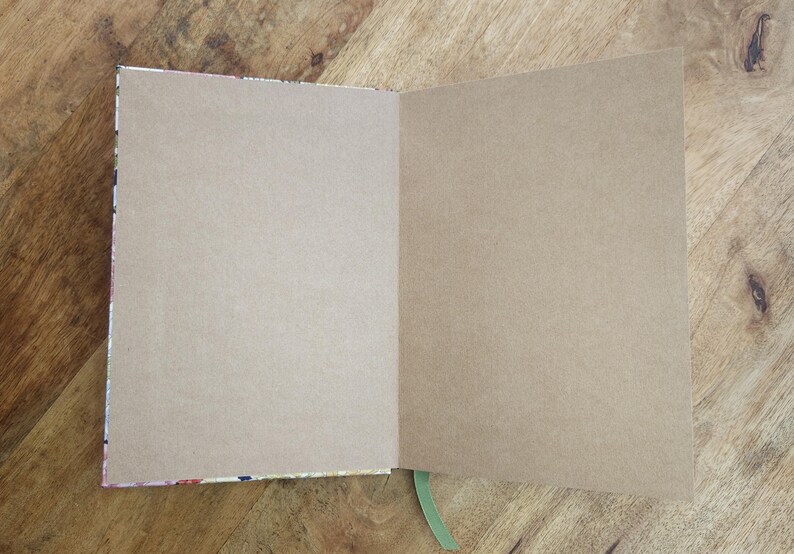 May include: An open notebook with blank, tan-colored pages. The notebook's binding has a colorful floral pattern. A green ribbon bookmark is visible. The notebook is on a wooden surface.