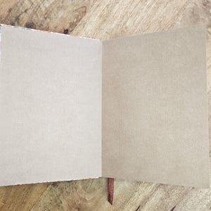 Puede incluir: Un cuaderno abierto con p&aacute;ginas en blanco de color marr&oacute;n claro. La encuadernaci&oacute;n del cuaderno es visible, con un borde estampado y un marcador de cinta marr&oacute;n. Las p&aacute;ginas son de color neutro y el cuaderno est&aacute; sobre una superficie de madera.