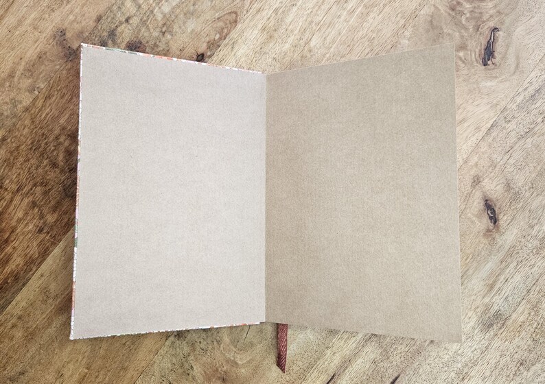 Puede incluir: Un cuaderno abierto con p&aacute;ginas en blanco de color marr&oacute;n claro. La encuadernaci&oacute;n del cuaderno es visible, con un borde estampado y un marcador de cinta marr&oacute;n. Las p&aacute;ginas son de color neutro y el cuaderno est&aacute; sobre una superficie de madera.