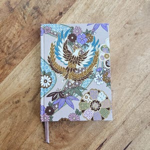 May include: A closed notebook with a decorative cover featuring a phoenix design in shades of brown, blue, and gold, surrounded by floral patterns. The notebook has a light pink ribbon bookmark.