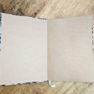 K&ouml;nnte beinhalten: Ein offenes Notizbuch mit leeren, hellbraunen Seiten. Der Einband hat ein Muster in Blau- und Wei&szlig;t&ouml;nen. Ein wei&szlig;es Lesezeichen aus Band ist sichtbar. Das Notizbuch liegt auf einer Holzoberfl&auml;che.